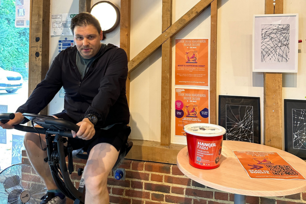 A man on a static bike in dark clothing, smiling at the camera. To the right of him is a table with a red donation bucket on and a poster flat. Behind the bucket on the wall are two orange posters.