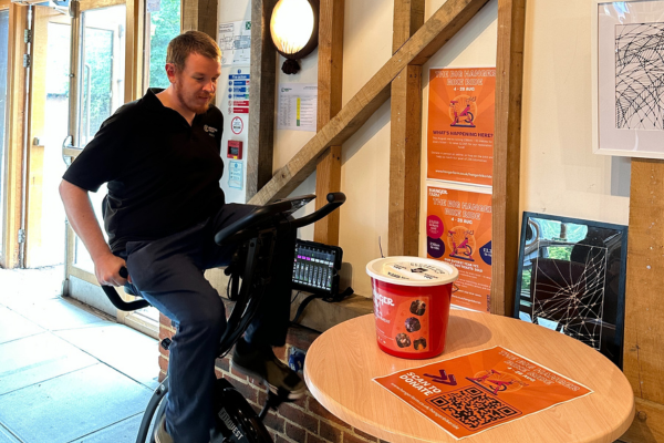 A man is sat on a static bike with a black tshirt and long black trousers. He is concentrating on cycling and looking down. To the right is a table with a red donation bucket and orange poster on top, behind is two orange info posters on the wall.