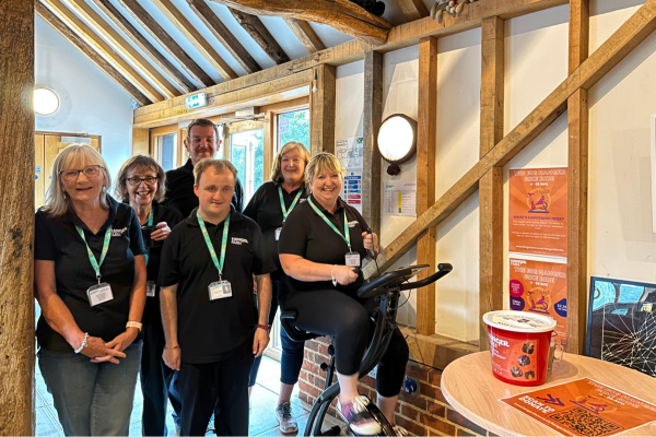 A group of 6 smiling volunteers stand next to a static bike with one volunteer on it. they are all wearing black tops and green lanyards. To the right is a table with a red donation bucket and orange poster on top, behind is two orange info posters on the wall.