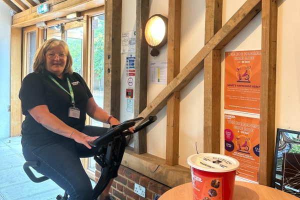A woman with grey shoulder length hair and glasses is smiling to the camera, sitting on a static bike. She is wearing jeans, a black tshirt and a green lanyard. To the right is a table with a red donation bucket and orange poster on top, behind is two orange info posters on the wall.