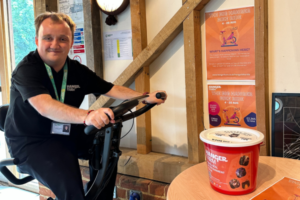 A man with short brown hair is smiling to the camera, sitting on a static bike. He is wearing black smart trousers, a black tshirt and a green lanyard. To the right is a table with a red donation bucket and orange poster on top, behind is two orange info posters on the wall. 