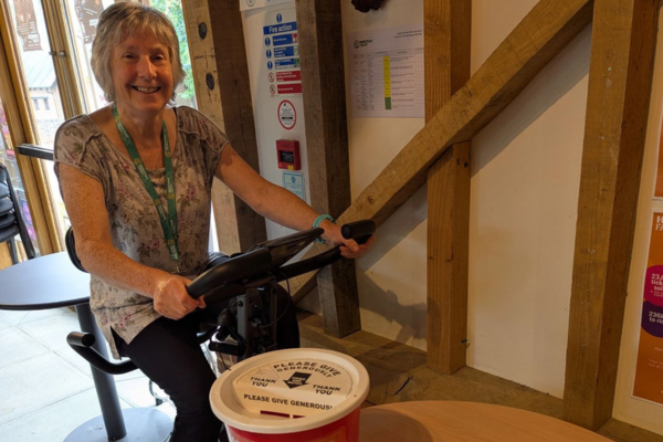 A woman with grey shoulder length hair and glasses is smiling to the camera, sitting on a static bike. She is wearing jeans, a black tshirt and a green lanyard. To the right is a table with a red donation bucket and orange poster on top, behind is two orange info posters on the wall.